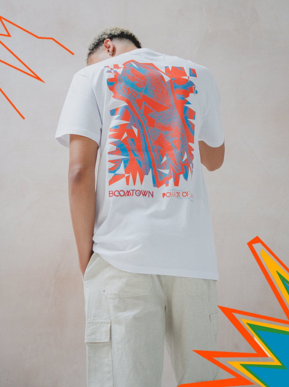A person faces away wearing the Uprising T-Shirt from Boomtown Fair Shop—a white organic cotton tee with a bold, abstract orange and blue graphic reading BOOMTOWN POWER OF—paired with light cargo pants against a neutral background with colorful shapes.