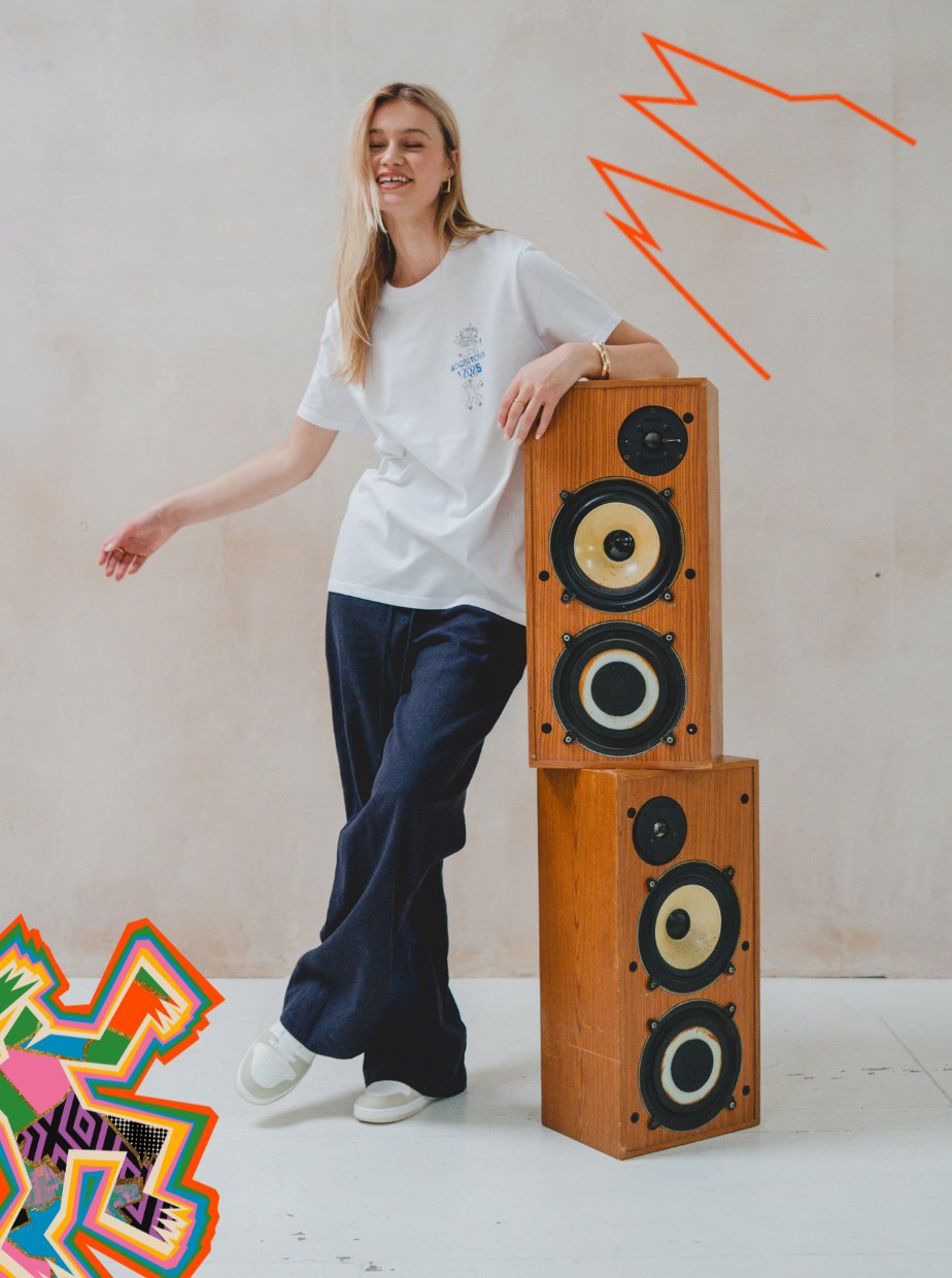 A smiling woman wearing the Boomtown Fair Shop’s Motion T-Shirt in organic cotton white and dark pants leans on stacked speakers, surrounded by colorful graphics and orange zigzags for a playful, creative vibe.