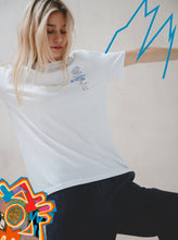 A young woman in the white Motion T-Shirt by Boomtown Fair Shop and dark pants poses with arms outstretched. Blue and red graphic doodles in the background add a playful, artistic flair with a hint of minimalist design.