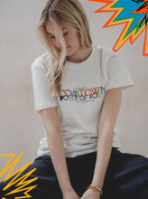 A young woman with long blonde hair looks down, wearing the Boomtown Fair Shop’s Power of Now: Line Up T-Shirt. Made from organic cotton, it features colorful DATOWN and POWER OF NOW text with abstract graphic accents in the image corners.