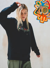 A person with long blonde hair wearing the Boomtown Fair Shop Power of Now: Line Up Hoodie, made from 100% organic soft cotton, and green pants stands indoors. A colorful abstract design appears in the top right corner.