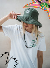 A person in a white T-shirt tilts down the 2025 Safari Hat Khaki, partially hiding their face. Colorful, abstract graphic shapes appear in the top right and bottom left corners of the image.