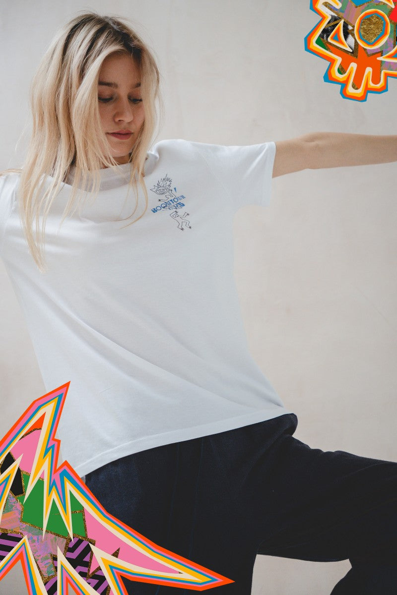A woman with long blonde hair wears the Motion T-Shirt by Boomtown Fair Shop—an organic cotton, minimalist white tee—paired with dark pants. She poses arms outstretched, framed by colorful abstract graphics in the image corners.