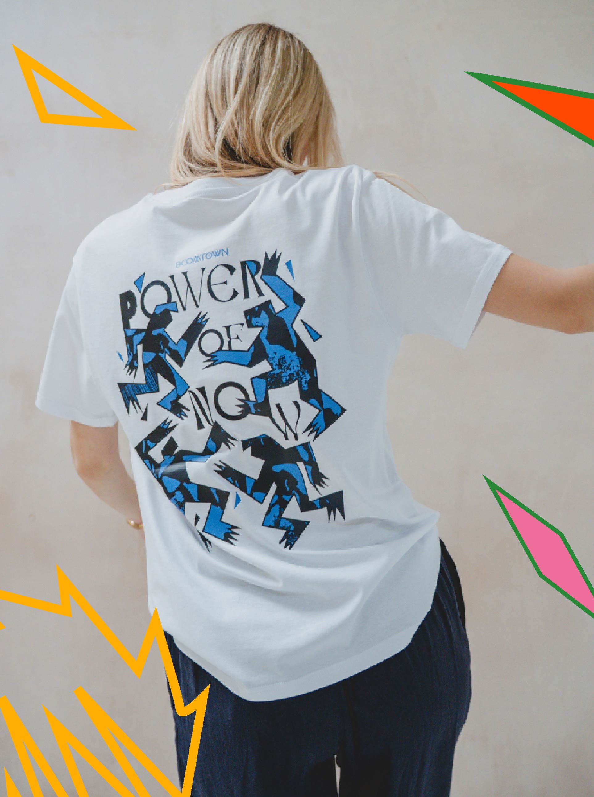 A person with long blonde hair faces away, wearing the Motion T-Shirt by Boomtown Fair Shop—a white organic cotton tee with a bold blue and black abstract minimalist design and Power of Now, accented by orange and pink graphic shapes at the border.