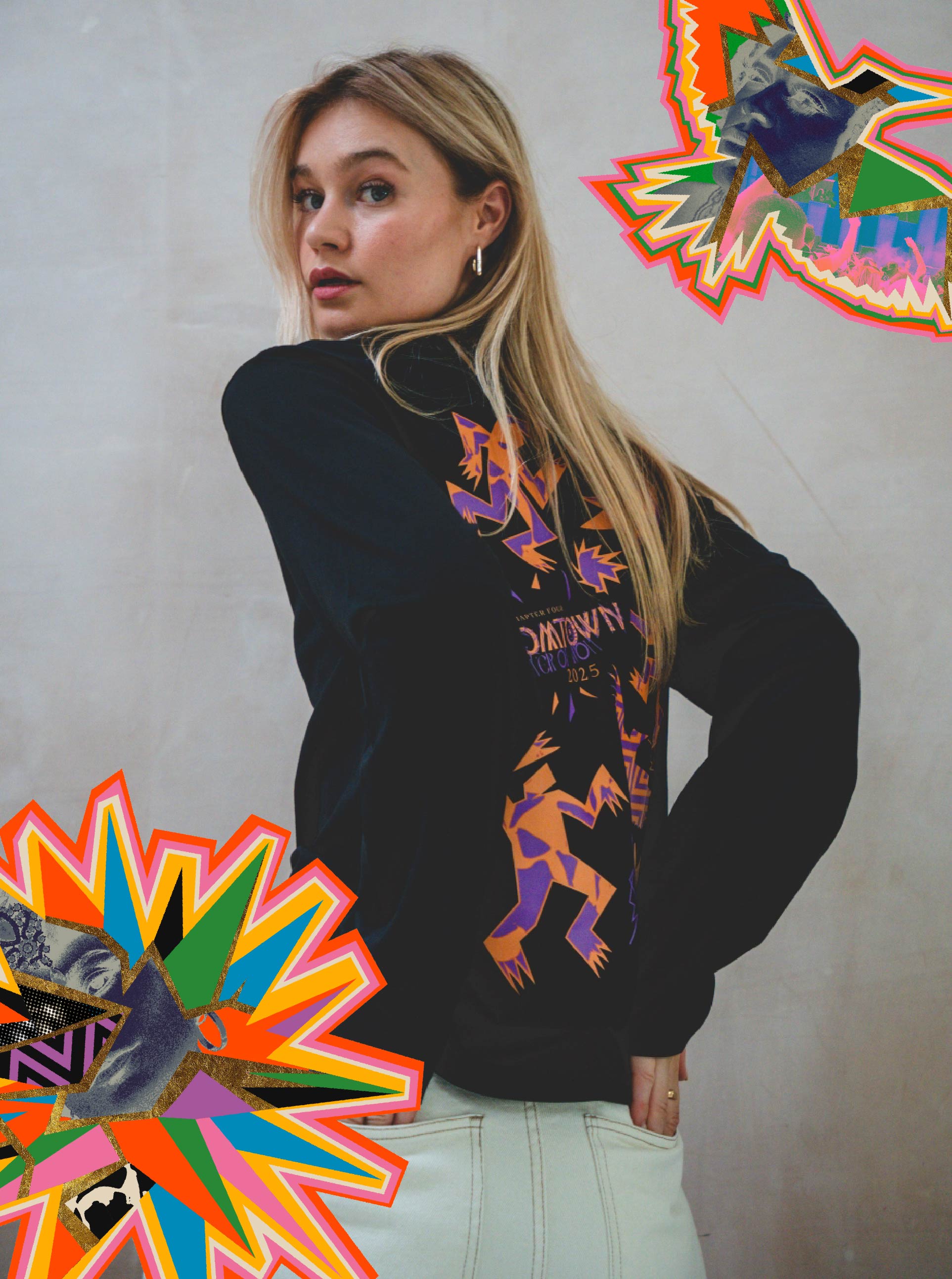 A woman with long blonde hair wears a Zephyr Long Sleeve black sweatshirt by Boomtown Fair Shop, featuring colorful graphic designs on the back. Bright abstract illustrations frame the image, making it perfect for festival season.