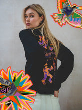 A woman with long blonde hair looks over her shoulder, wearing the Boomtown Fair Shop Power of Now Icon Graphic Long Sleeve T-Shirt, featuring bold, colorful abstract designs and vibrant starburst patterns—perfect for festival season.