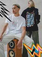 A man wears the Boomtown Fair Shops Power of Now Chroma T-Shirt, crafted from organic soft cotton, as he sits on a speaker; beside him stands a woman in a black hoodie with colorful text. Bold shapes highlight the Power of Now theme.