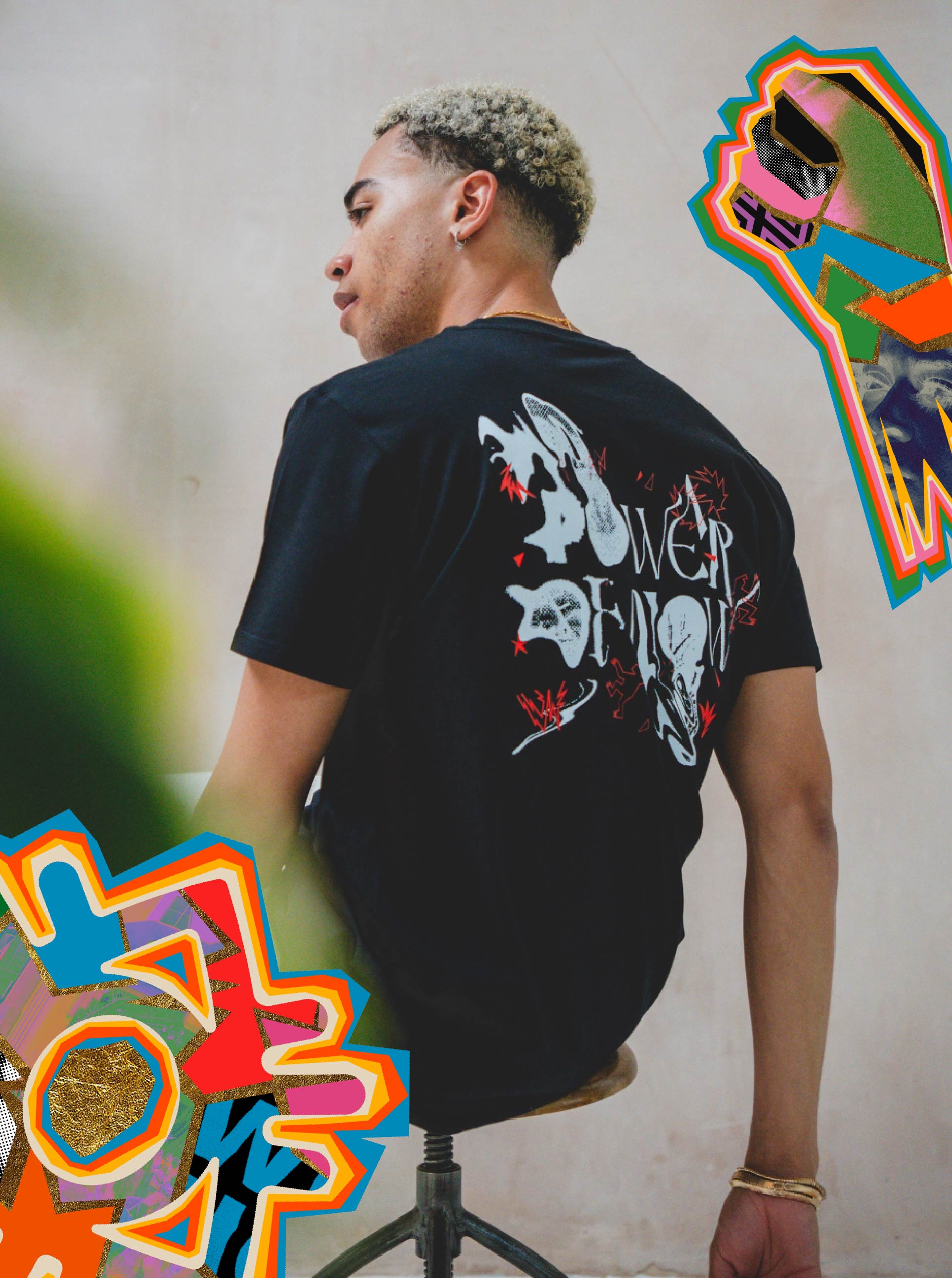 A young man with short, curly, blond-tipped hair sits on a stool wearing the Boomtown Fair Shops Power of Now Mono Graphic T-Shirt. Colorful abstract designs in the foreground and top right add vibrancy to the minimalist style.