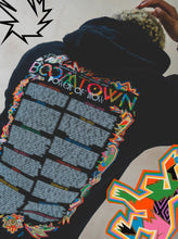 A person wears the 2025 Power of Now: Line Up Hoodie, featuring bold, abstract graphics and the phrase POWER OF NOW on the back. Colorful shapes and patterns frame the image’s borders.