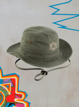 The 2025 Safari Hat Khaki features ventilation eyelets, an adjustable strap, and a beige abstract eye symbol on the side—set against a light background with colorful geometric designs, making it an ideal festival accessory.
