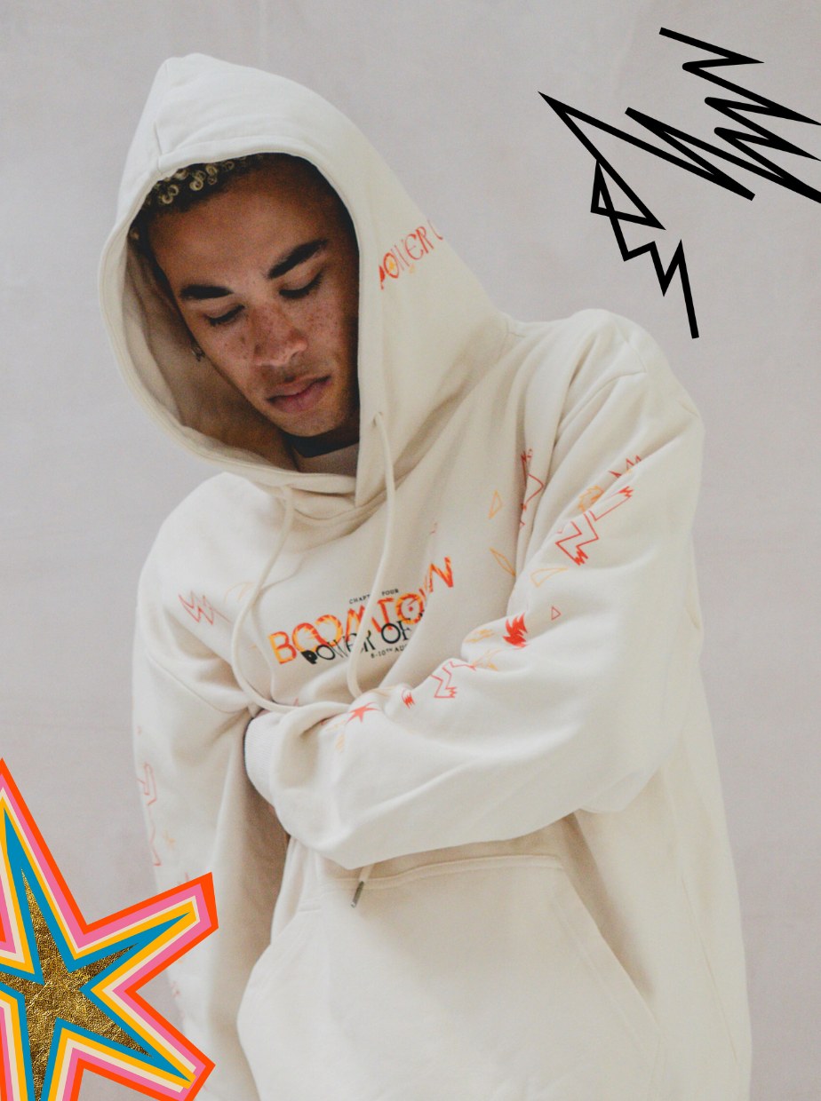 A person in the 2025 Power of Now: Limited Edition Emberkind Hoodie, made from organic cotton, stands with their head slightly bowed. The cream hoodie features orange designs and graphic black and orange star and zigzag accents.