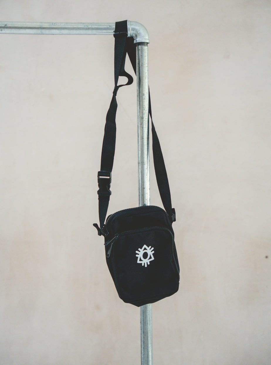 The Boomtown Fair Shop Cross Body Bag, featuring an adjustable strap and a white abstract symbol on black, hangs from a metal rack against a beige background.