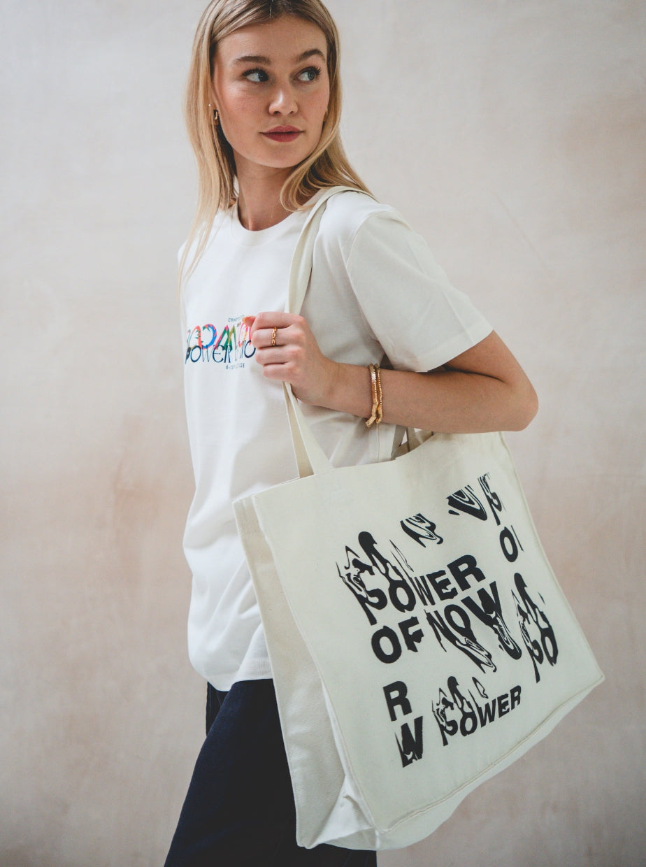 A woman with long blonde hair wears a white t-shirt and dark pants, holding the Boomtown Fair Shop Canvas Tote Bag with bold black text and illustrations. She gazes to the side against a neutral background.