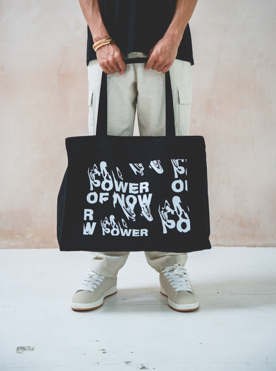 A person stands holding the Black Tote Bag from Boomtown Fair Shop, featuring bold white distorted text reading POWER OF NOW, styled with light cargo pants, white sneakers, and a black long-sleeve shirt against a plain background.